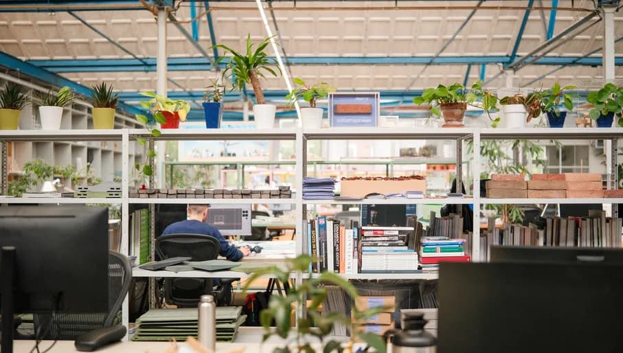 Man working in an office at a computer, with shelves holding plants, books and circular building materials. From the FRONT offices.