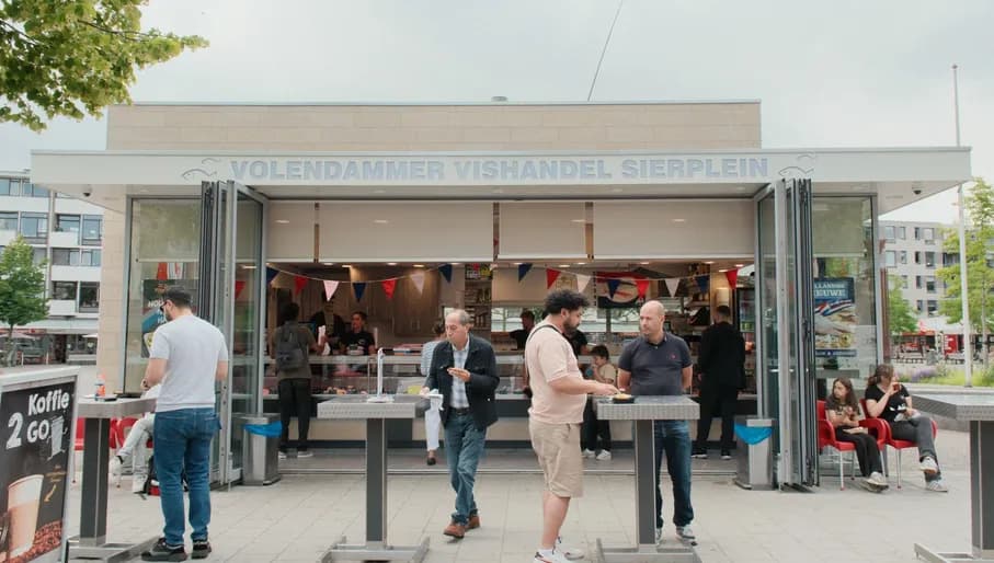 The photo was taken at Sierplein 86 in Nieuw-West, and shows herring stall Volendam Stroek Sierplein. It captures people eating at the tables in front of the stall or waiting to be served.