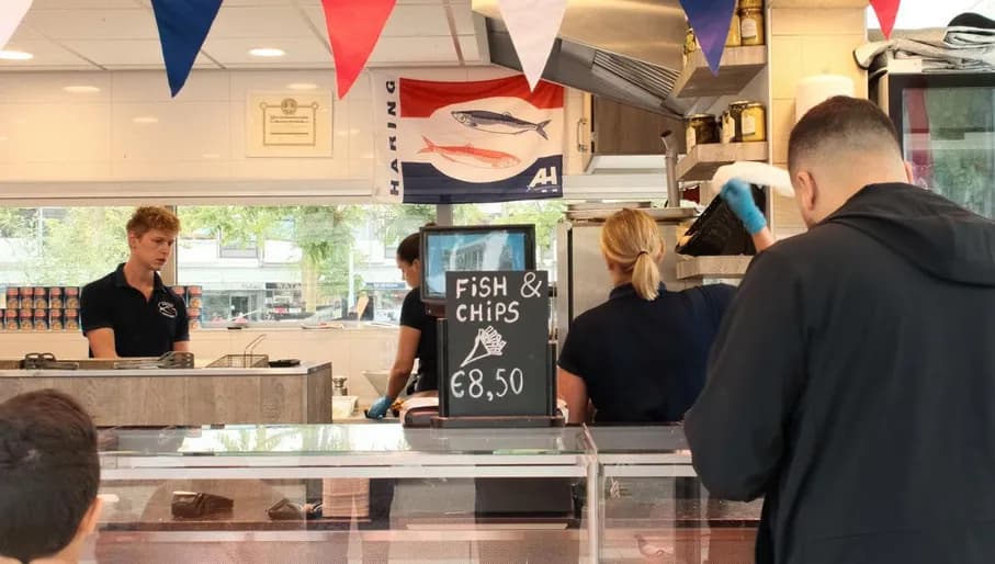 The photo was taken at Sierplein 86 in Nieuw-West, and shows herring stall Volendam Stroek Sierplein. It captures a man and a boy waiting to be served. Decorations in the colours of the Dutch flag, a sign with Fish & Chips and fried food behind the glass counter are also visible.