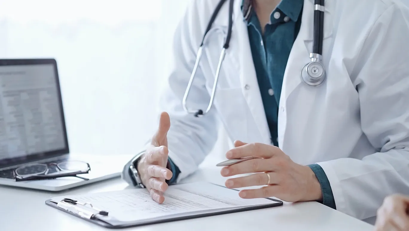 Doctor and a patient. The physician, wearing a white medical coat over a green shirt, is gesturing with his hands during a consultation in the clinic. Medicine