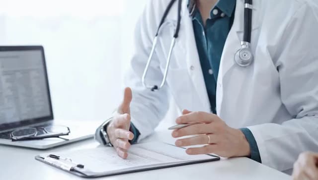 Doctor and a patient. The physician, wearing a white medical coat over a green shirt, is gesturing with his hands during a consultation in the clinic. Medicine