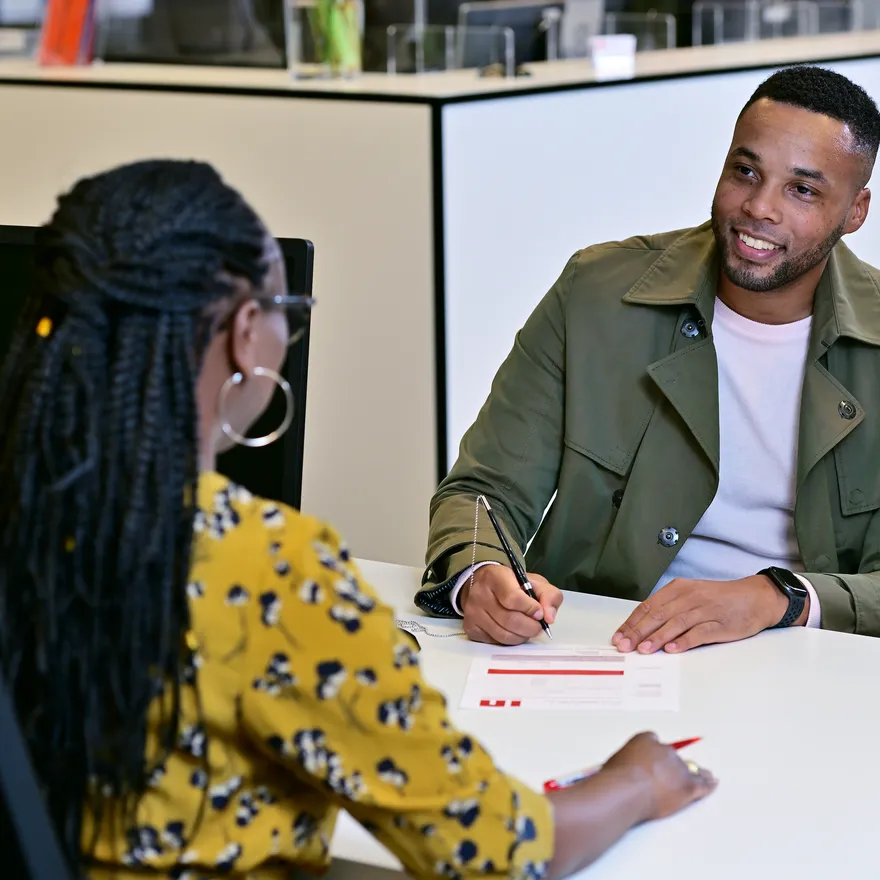 Man sitting at the desk in the INAmsterdam offices, seen from the front, talking to employee, seen from the back