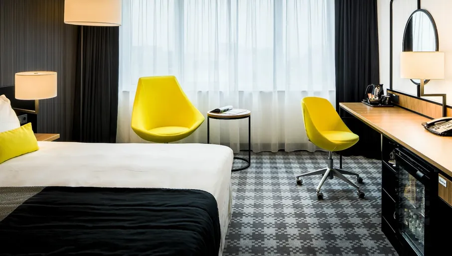 Interior of Radisson Blu Hotel Amsterdam Airport room, with bed, desk mini fridge, yellow chairs and window with blinds drawn.