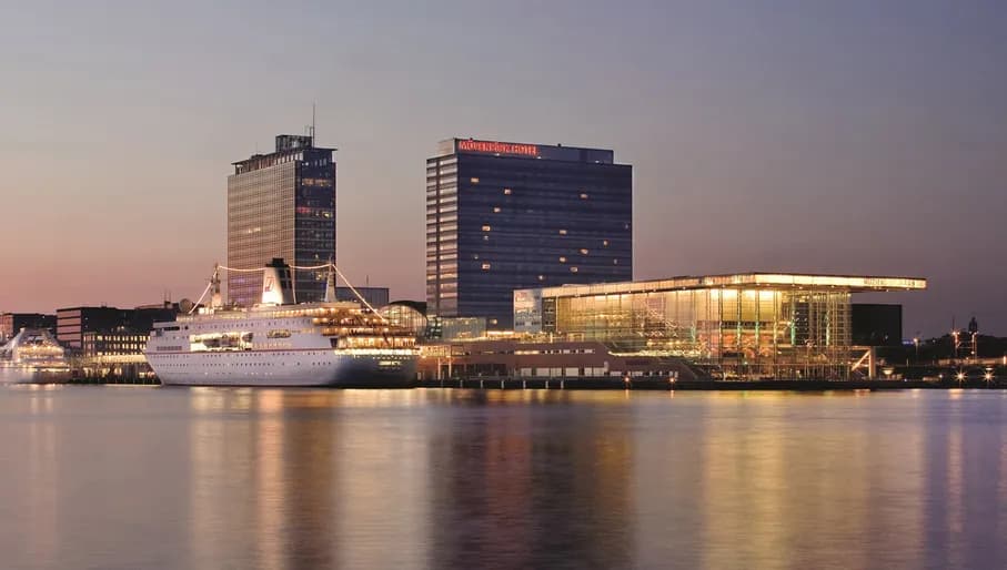 Long exterior shot of Mövenpick Hotel Amsterdam City Centre at dusk, from water of IJ River, with ferry and Muziekgebouw aan 't IJ in the foreground.