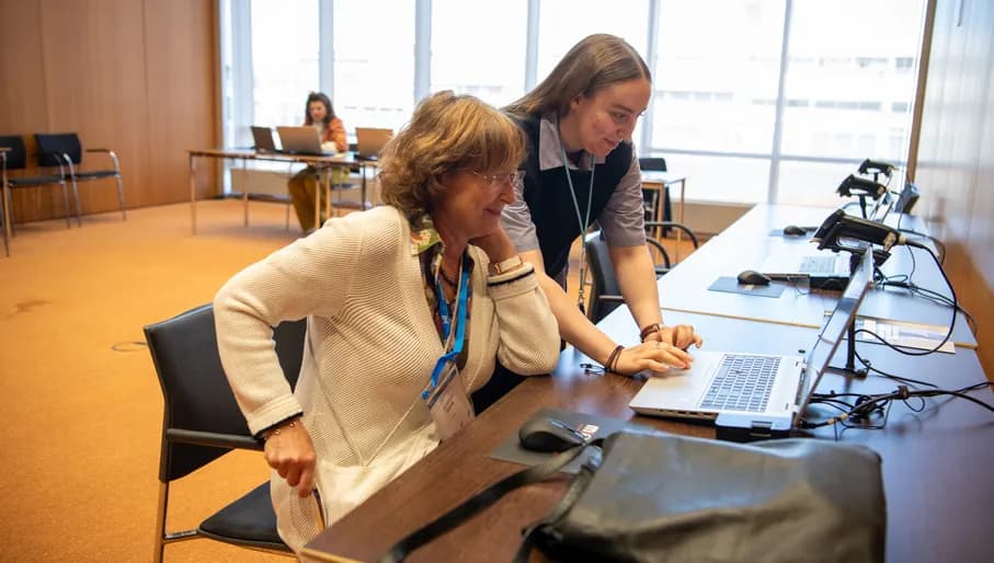 Two women collaborate over a laptop in a medical, formal setting.