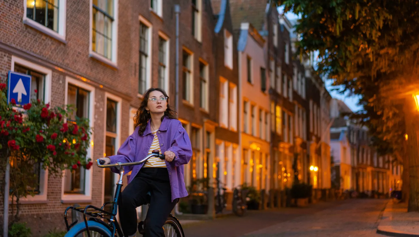 Young Caucasian woman in purple coat riding bicycle in the city at dusk