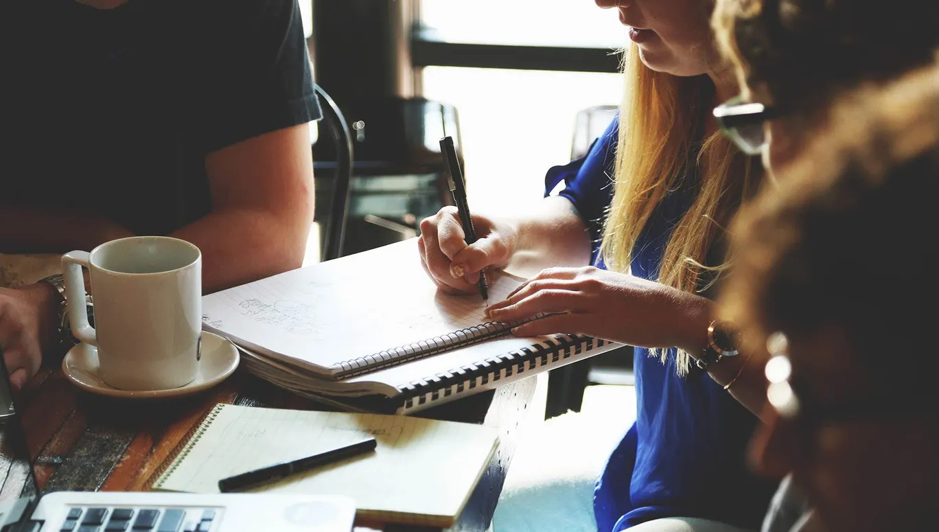 Closeup of hands taking notes on pad of paper next to coffee cup at desk during meeting.